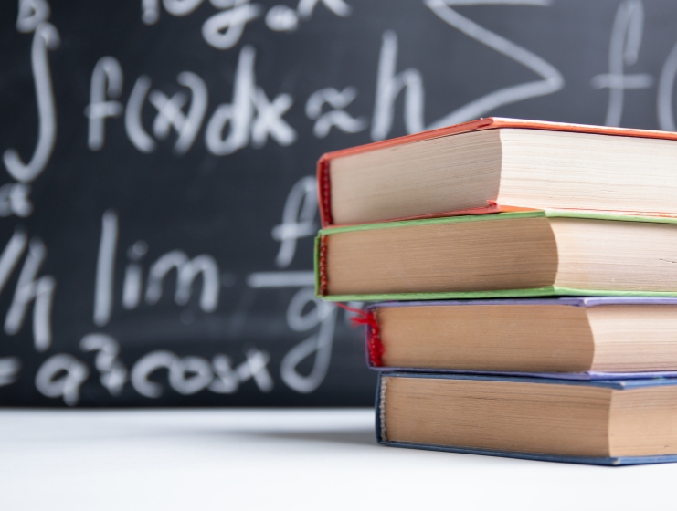 A stack of textbooks in front of a chalkboard with math formulas, representing books to master advanced mathematics