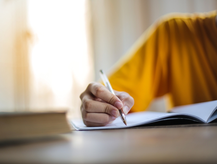 A person in a yellow shirt writing notes in a notebook at a desk with a book nearby