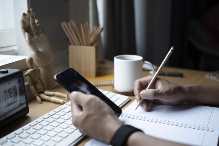 Cropped shot male using mobile phone and making note on notebook.